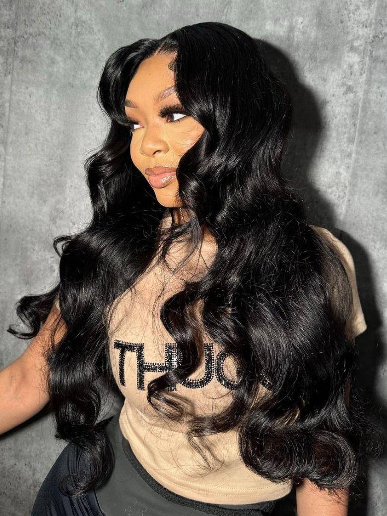 Woman with long black wavy wig wearing a beige graphic t-shirt posing against gray backdrop