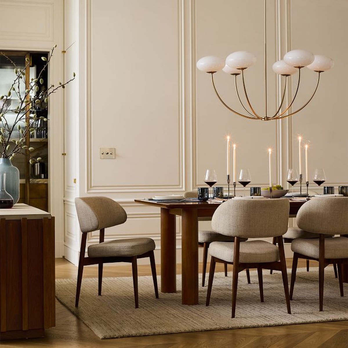 Elegant dining room with beige paneled walls, wooden table, neutral upholstered chairs, and decorative chandelier