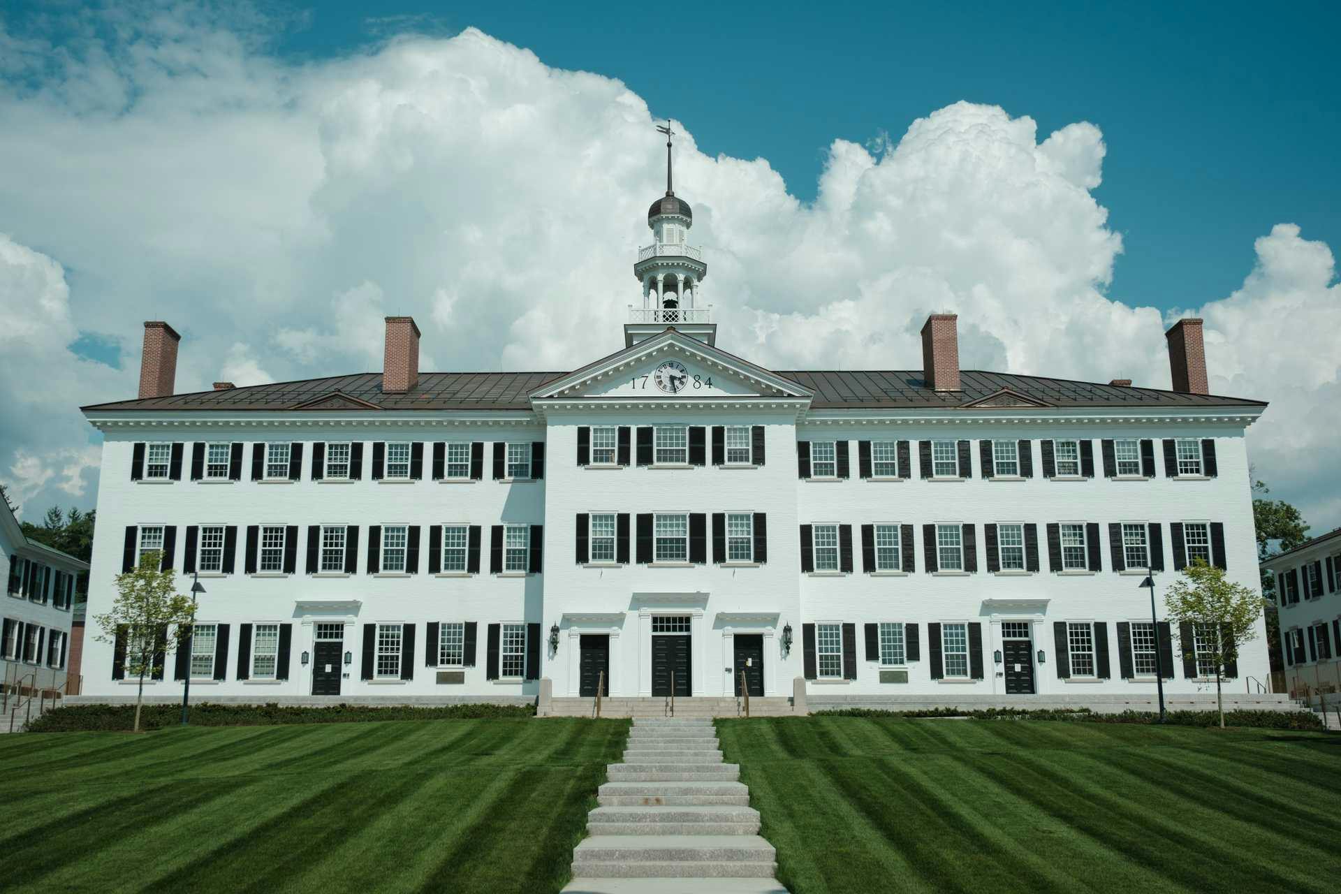 White colonial-style building with black shutters, cupola, and manicured lawn under blue sky with clouds
