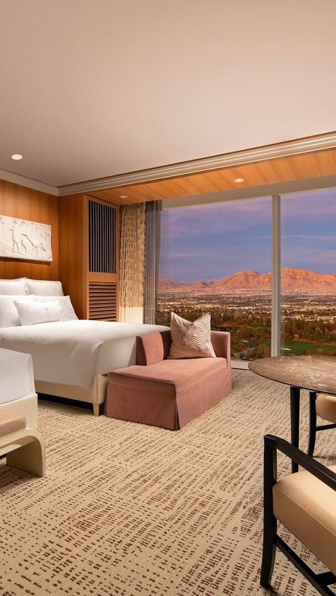 Luxury hotel bedroom with wood paneling, white bed, pink accent furniture, and floor-to-ceiling windows with desert mountain views