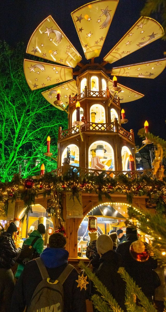 Illuminated three-tier wooden Christmas pyramid with star-patterned sails and crowds gathered around at night market
