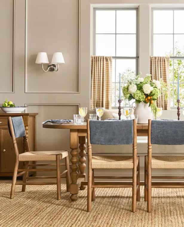 Dining room with wooden table, upholstered chairs, large windows with plaid curtains, and fresh flowers in vase