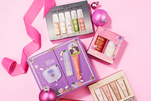 Holiday beauty gift sets with body mist, fragrance kit, and skincare products surrounded by pink ribbon and ornaments