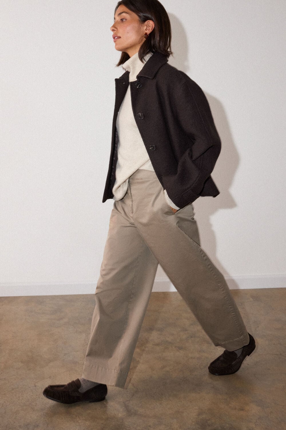 Woman with shoulder-length dark hair wearing black blazer, cream sweater, and beige trousers