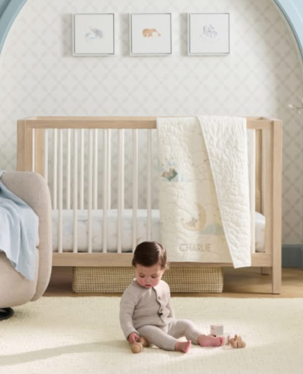Nursery with natural wood crib, cream bedding, and framed animal artwork above, baby sitting on beige floor