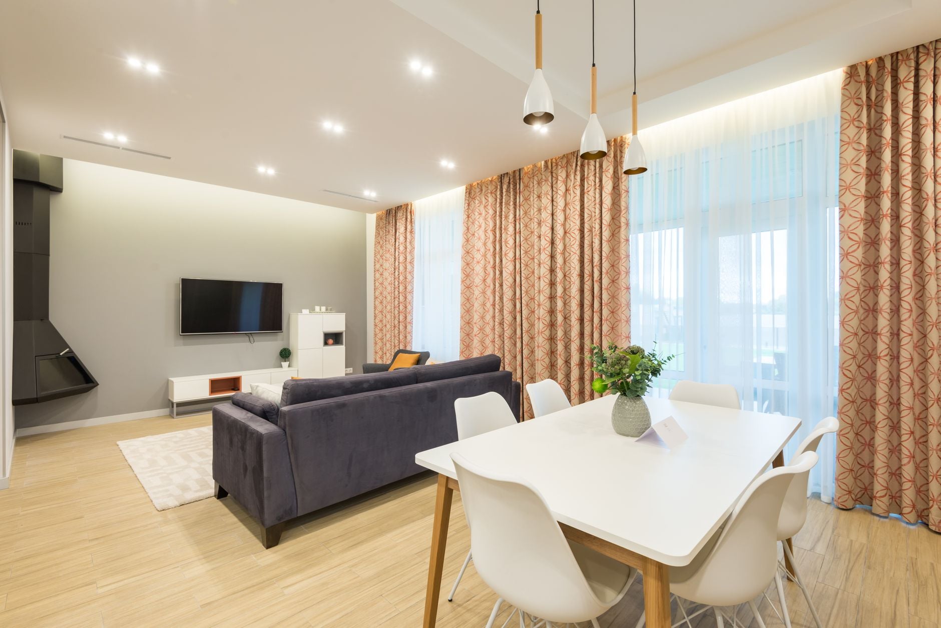 Modern living and dining area with dark leather sofa, white dining table, patterned curtains, and pendant lights
