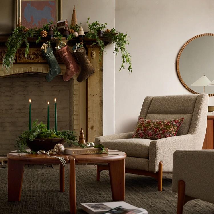 Festive living room with decorated fireplace mantel, stockings, garland, gray wingback chair, and round wooden coffee table