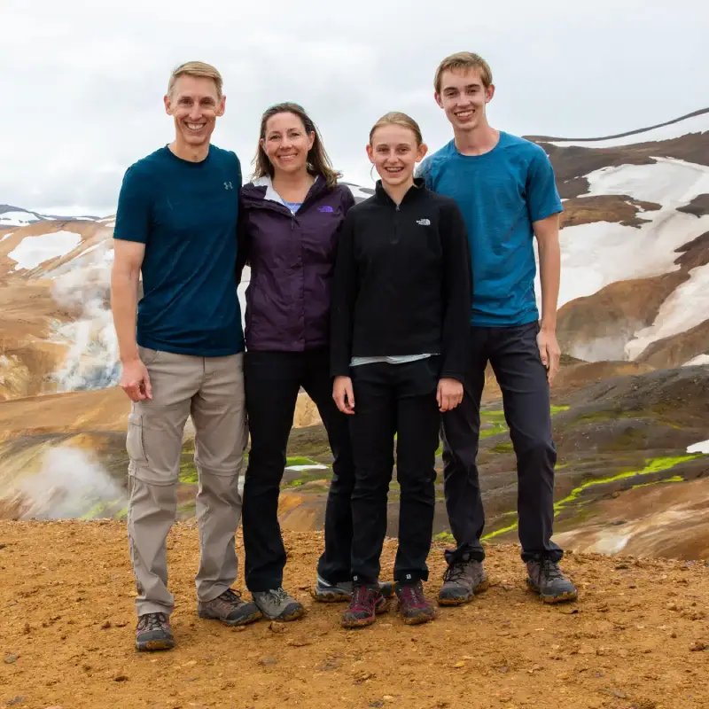 Four people smiling at camera in hiking gear with geothermal landscape of colorful hot springs and snow-capped mountains behind them