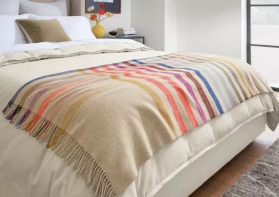 Colorful striped bed blanket with fringe draped over neutral bedding with olive pillow