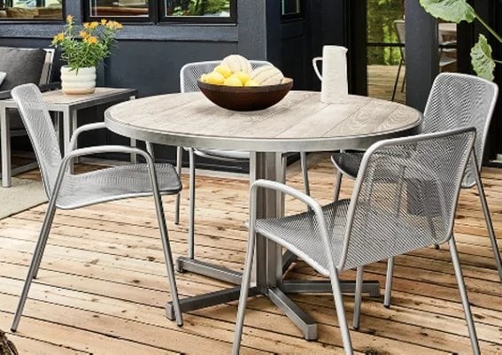 Round light wood outdoor dining table with four metallic mesh chairs on wooden deck