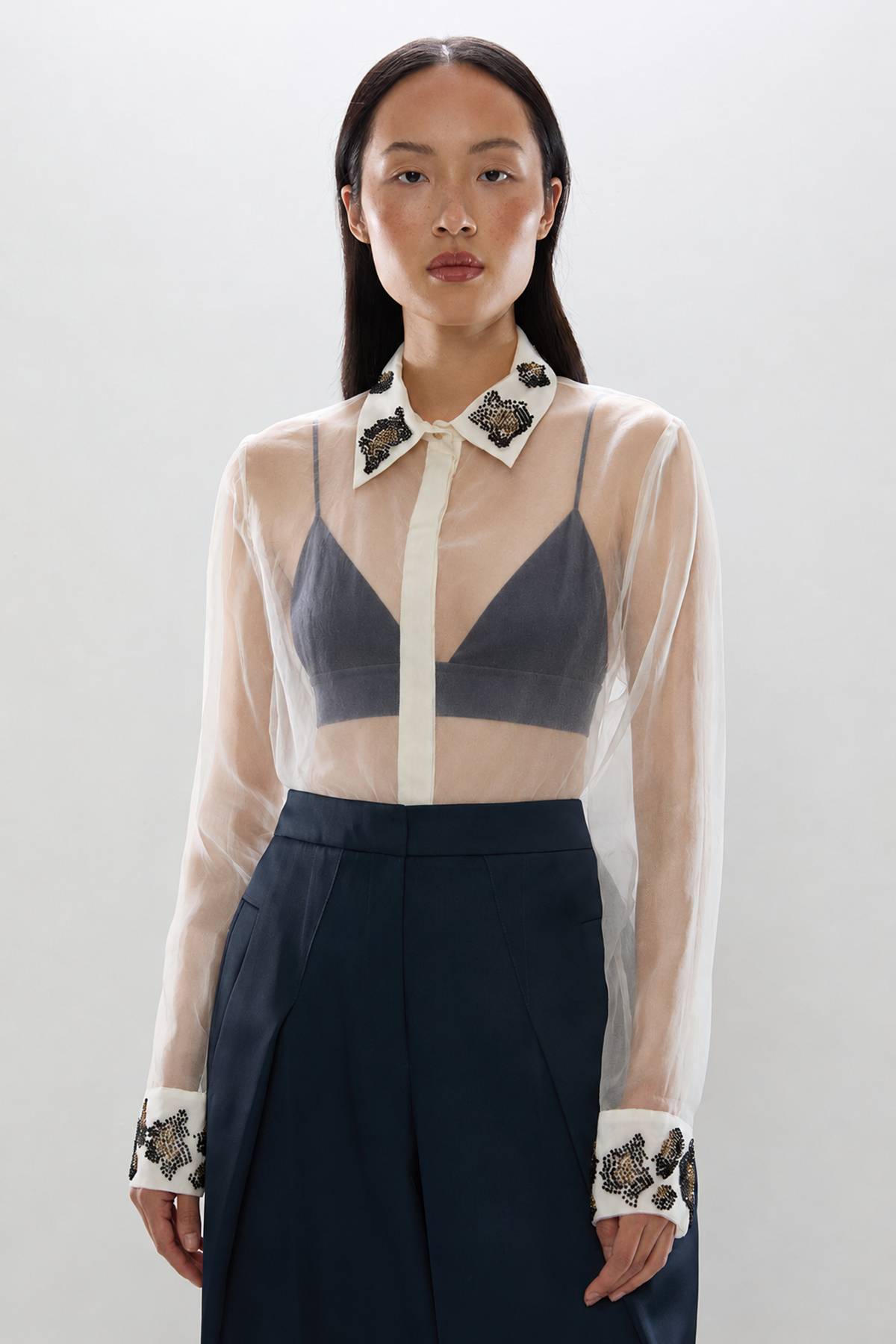 Woman wearing cream sheer blouse with beaded collar and cuffs over black bra and navy skirt