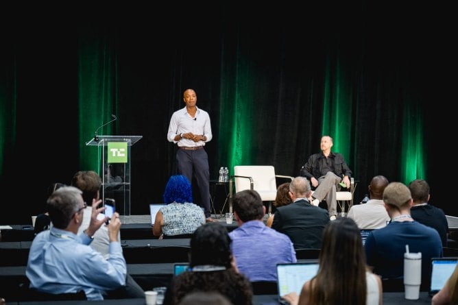 Man speaking at a conference podium with green lighting and audience members visible