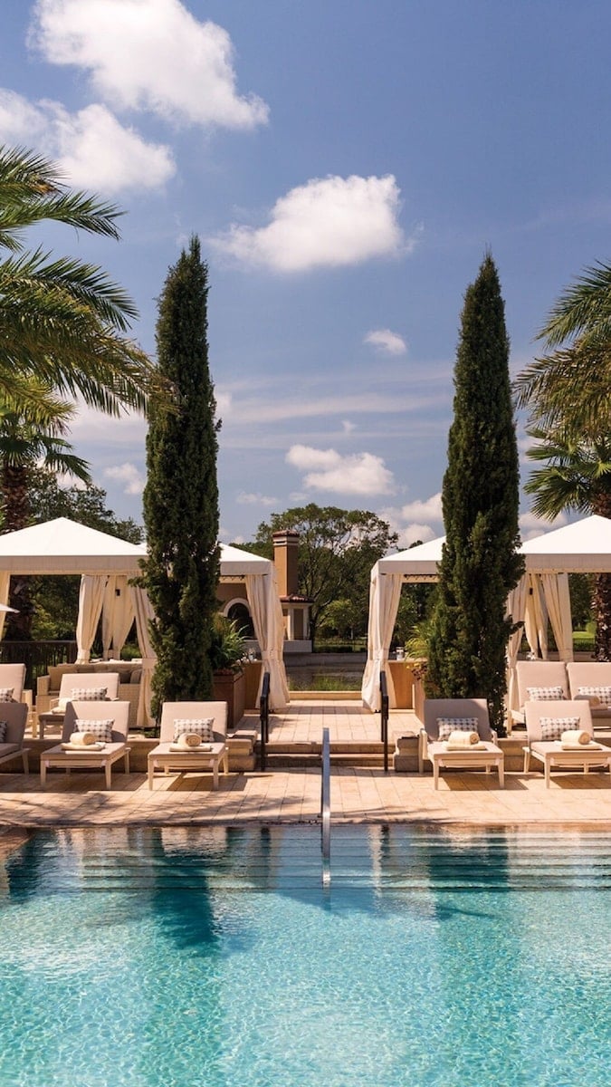 Upscale resort pool area with tall cypress trees, white loungers, cabanas, and pristine blue water under cloudy sky