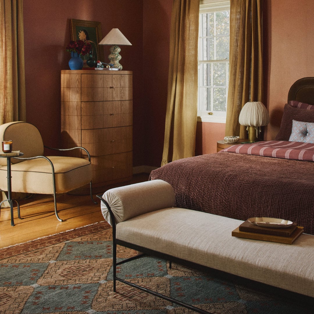 Warm-toned bedroom with burgundy walls, golden curtains, wooden dresser, upholstered bench, and bed with textured brown bedding