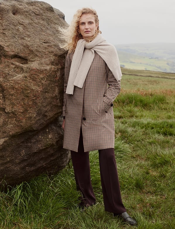 Woman with curly blonde hair wearing a brown gingham coat and cream scarf standing against a large boulder in moorland