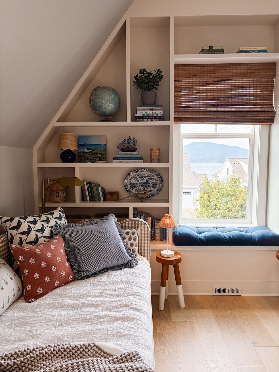 Bedroom with sloped ceiling, built-in shelving displaying books and decorative objects, bed with patterned pillows, and window seat overlooking water