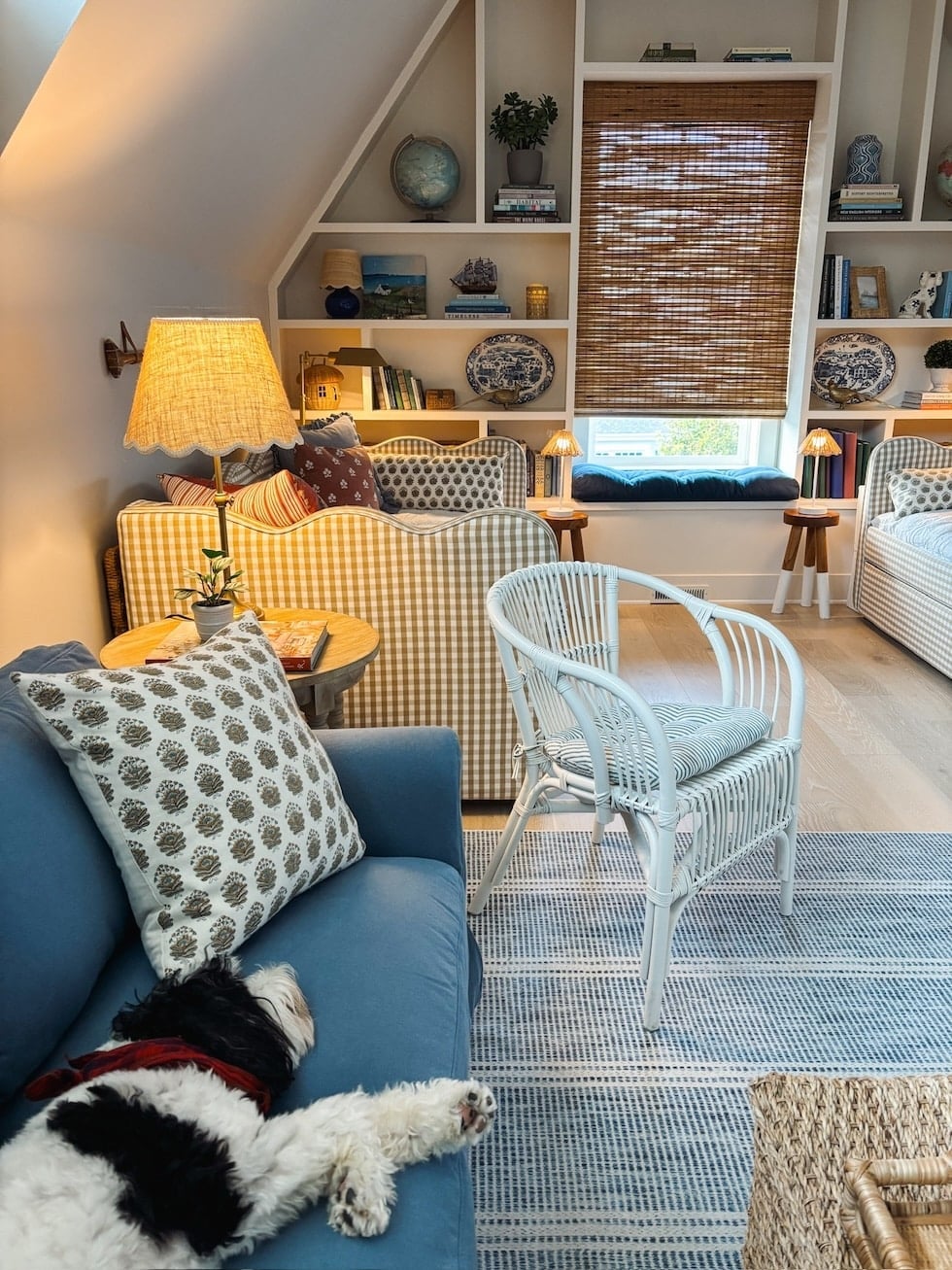 Attic bedroom lounge with yellow-checked upholstered sofa, blue armchair, white wicker chair, built-in shelving with books and decor, and roman shades
