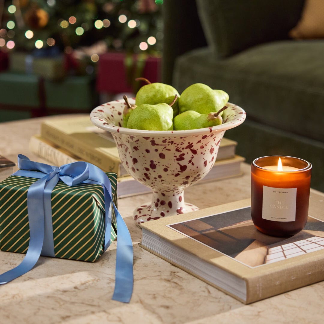 Marble table surface styled with wrapped gift, potted fruit bowl, lit candle, and magazines with holiday decorations blurred in background