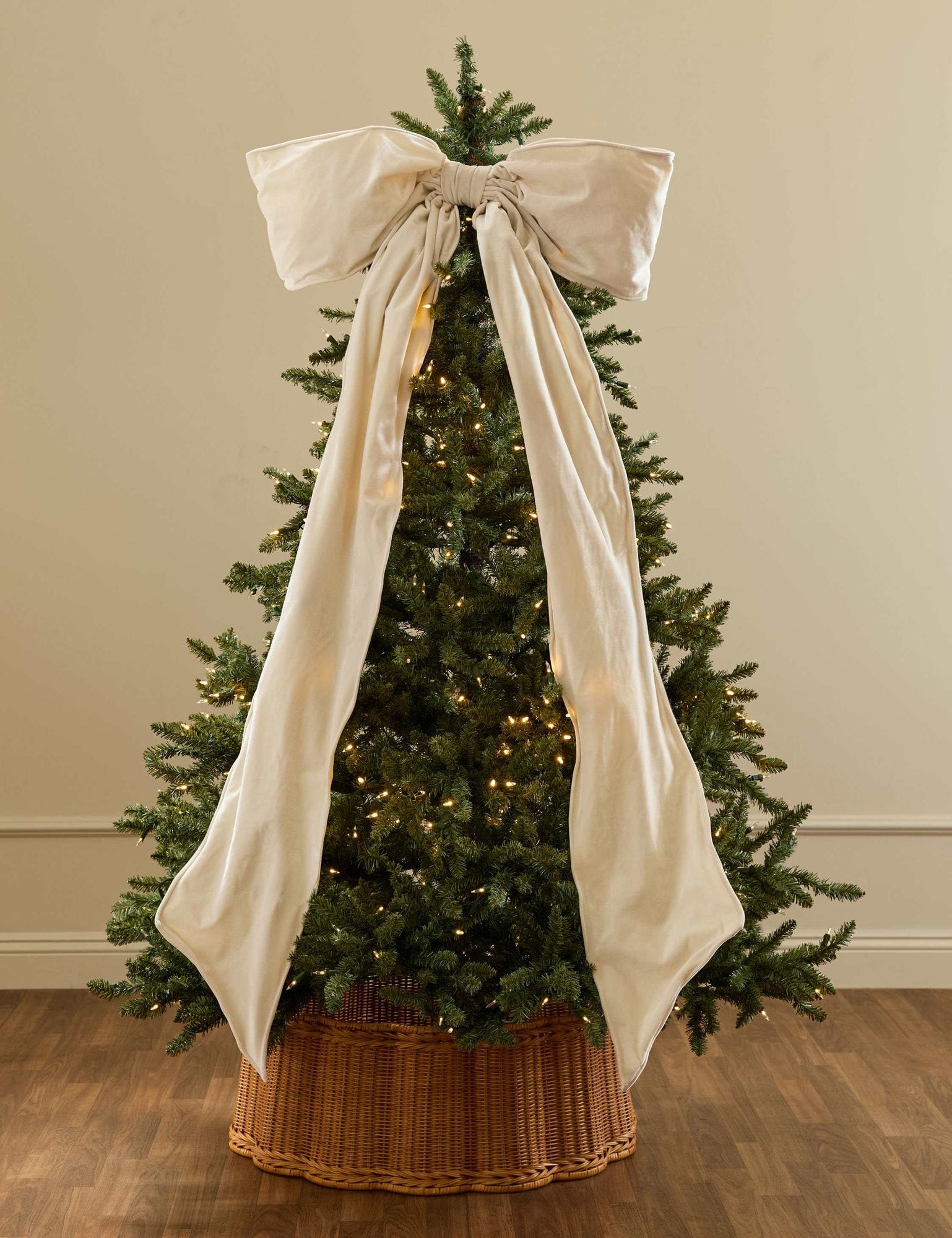 Christmas tree decorated with warm white lights in woven basket, topped with cream-colored bow and matching fabric ribbon draping