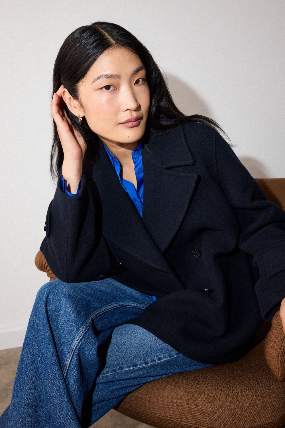 Woman with long black hair sitting on brown chair wearing navy blazer, blue shirt, and blue jeans