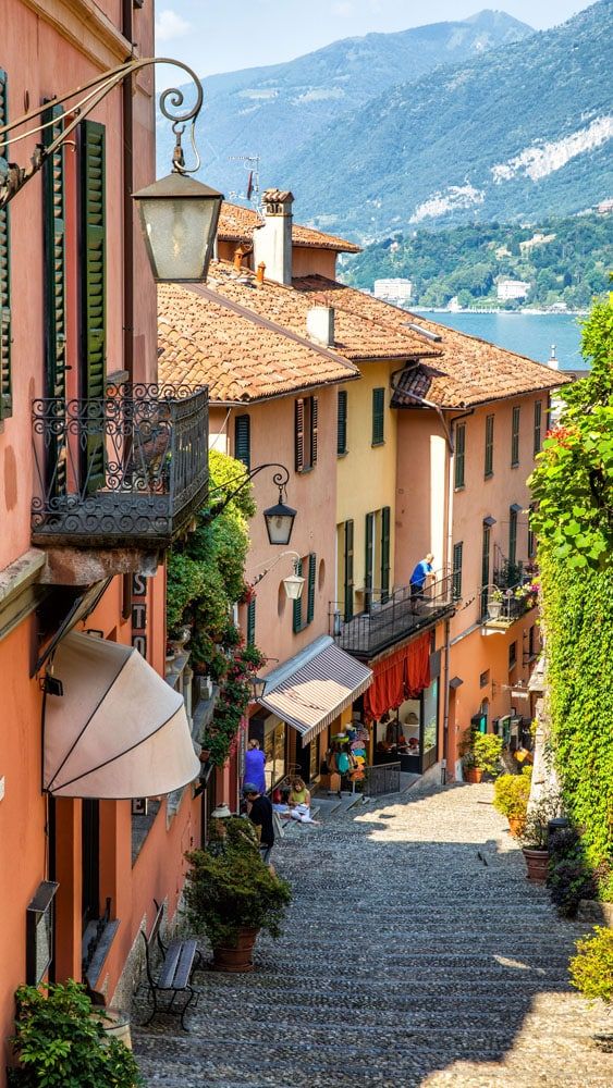 Steep narrow street lined with colorful Italian buildings, mountain, and lake visible in distant background