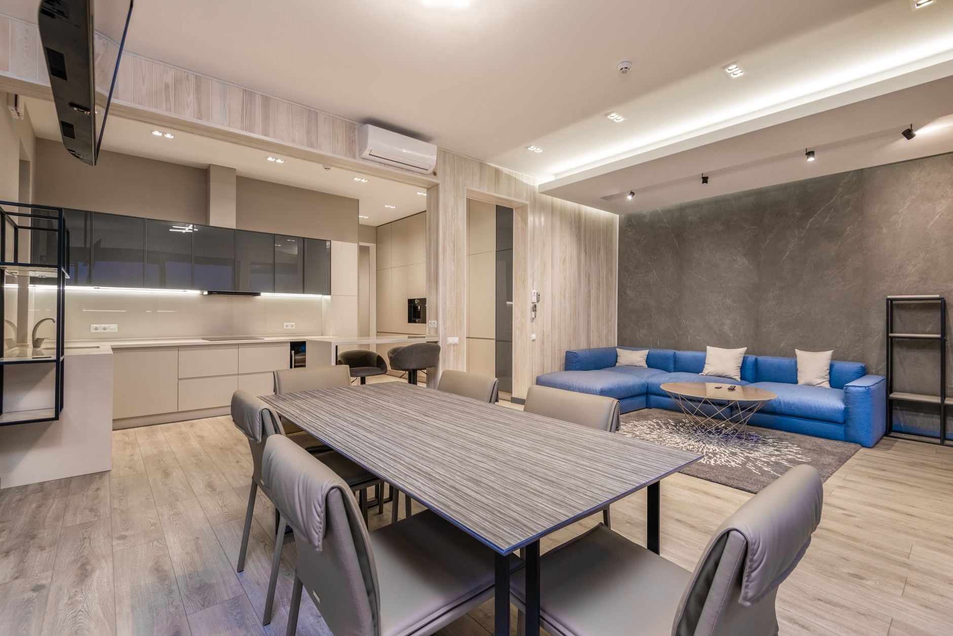 Contemporary open-plan apartment with blue sectional sofa, striped dining table, kitchen, and textured accent wall