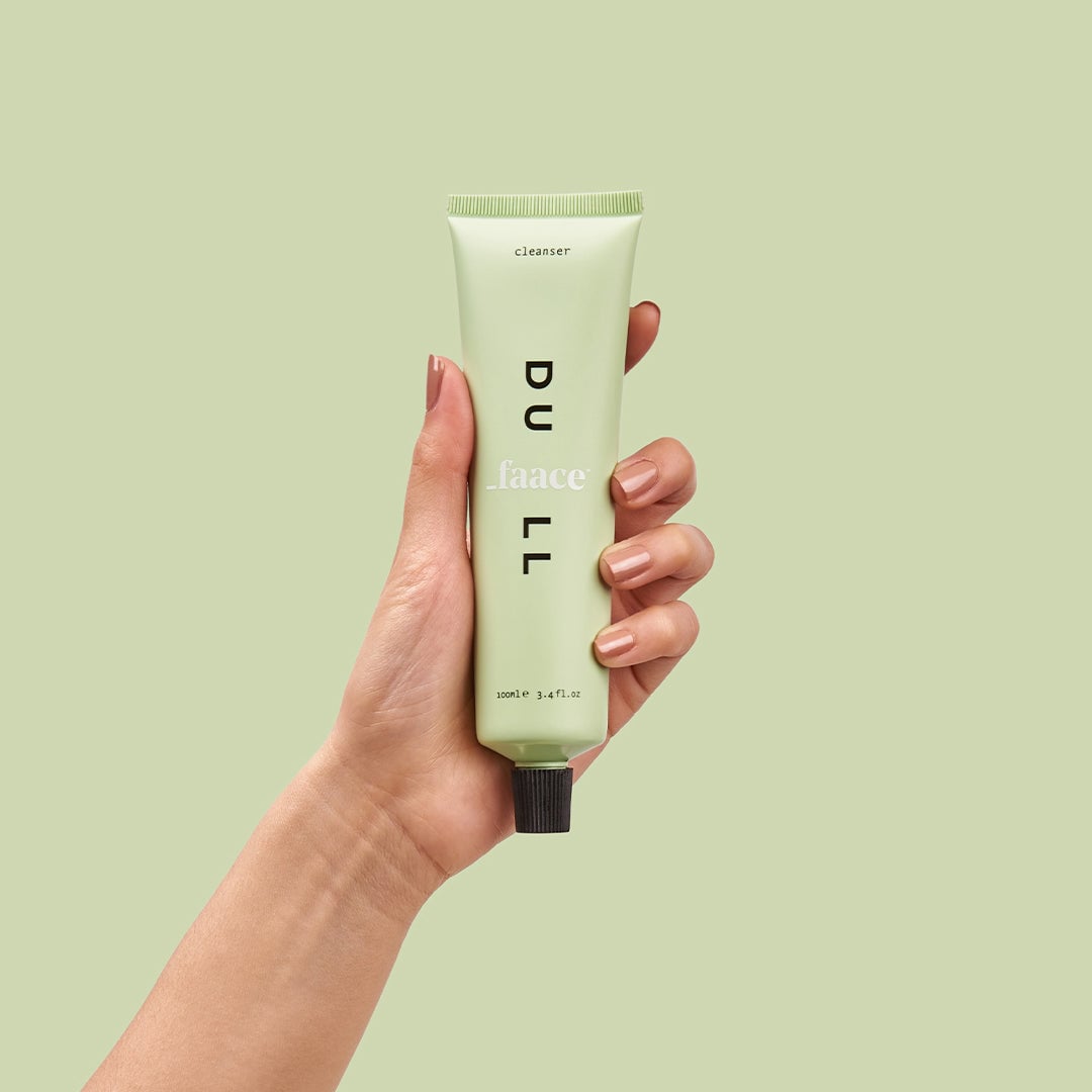 Hand holding pale green facial cleanser tube against matching background