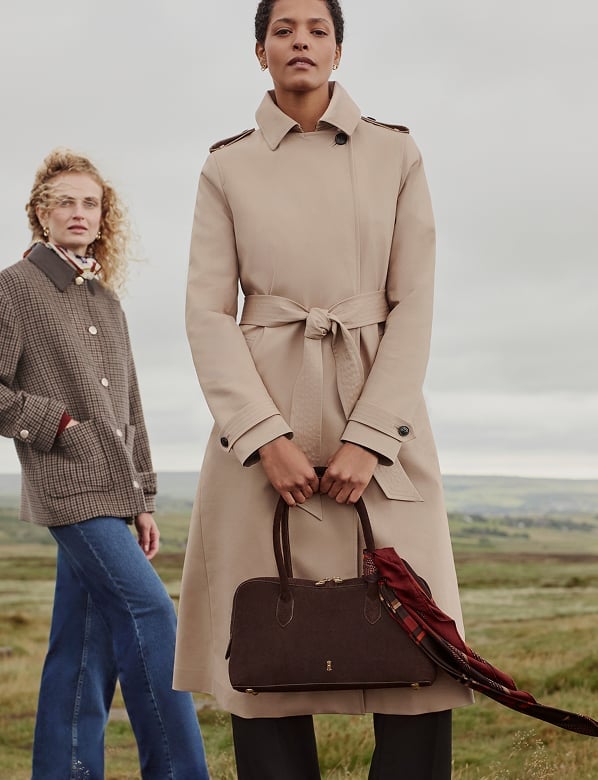 Two women on a hillside, one wearing a brown gingham jacket with jeans, the other in a beige trench coat holding a brown handbag
