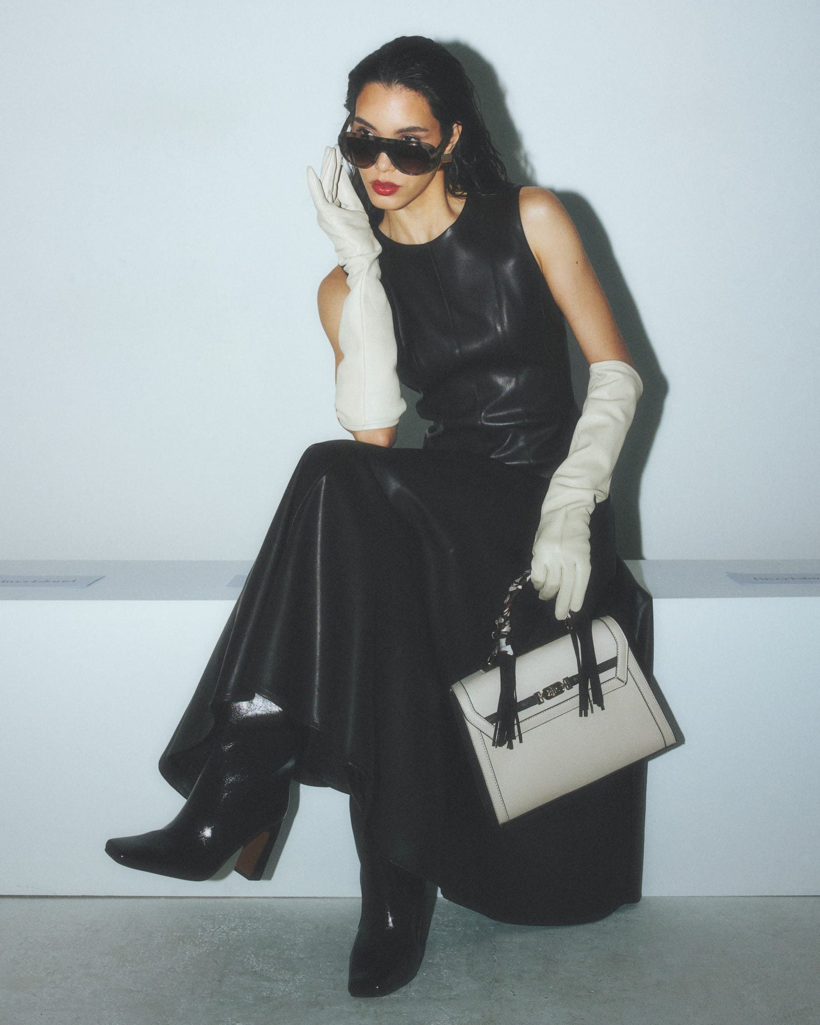 Woman wearing black sleeveless dress with white gloves, sunglasses and cream handbag