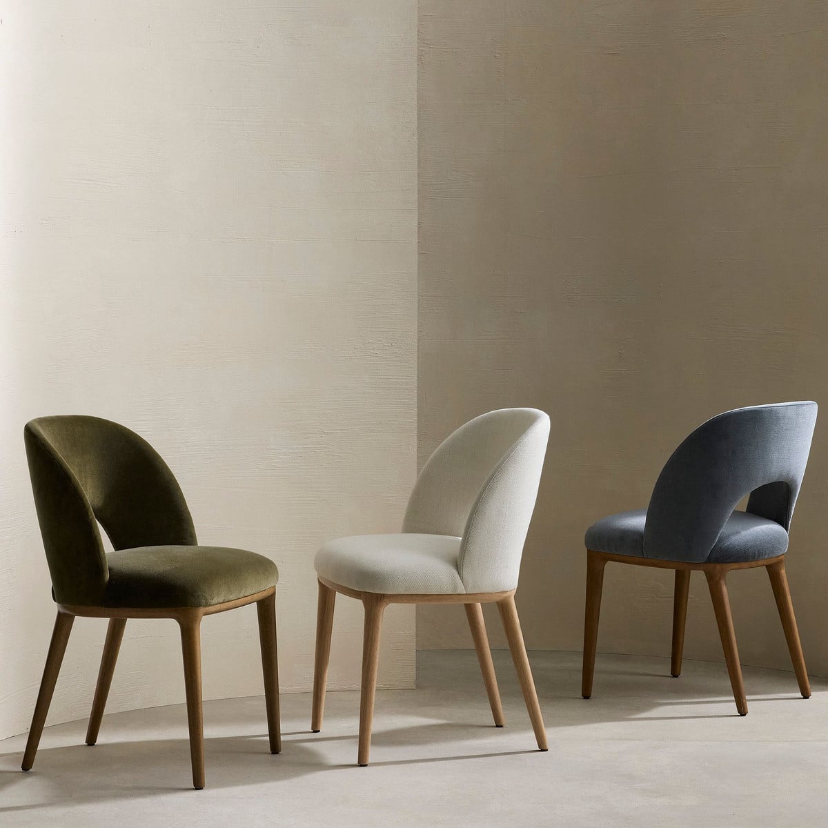 Three modern dining chairs with curved backs in olive, cream, and navy upholstery with natural wood legs
