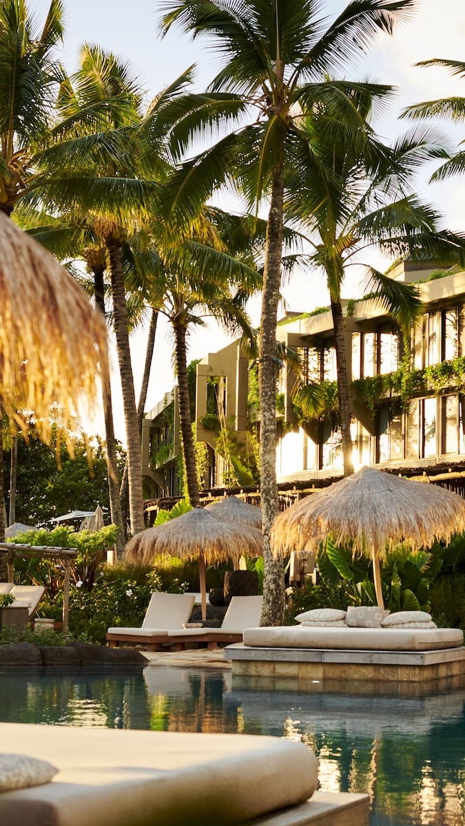 Thatched umbrellas and white loungers by a calm pool with tall palm trees and modern architecture in background