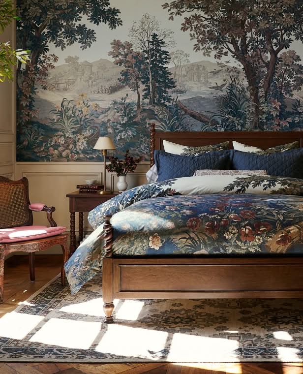 Bedroom with ornate toile wallpaper mural, wooden bed with blue patterned bedding, and antique nightstand with lamp