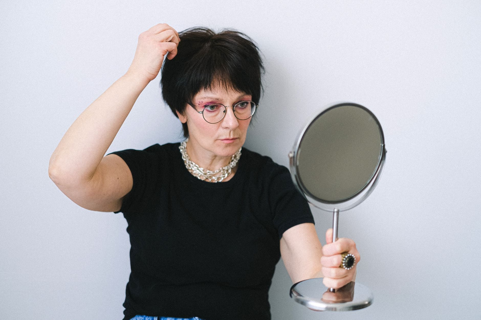 Person with short dark hair and glasses examining their hair in a hand mirror