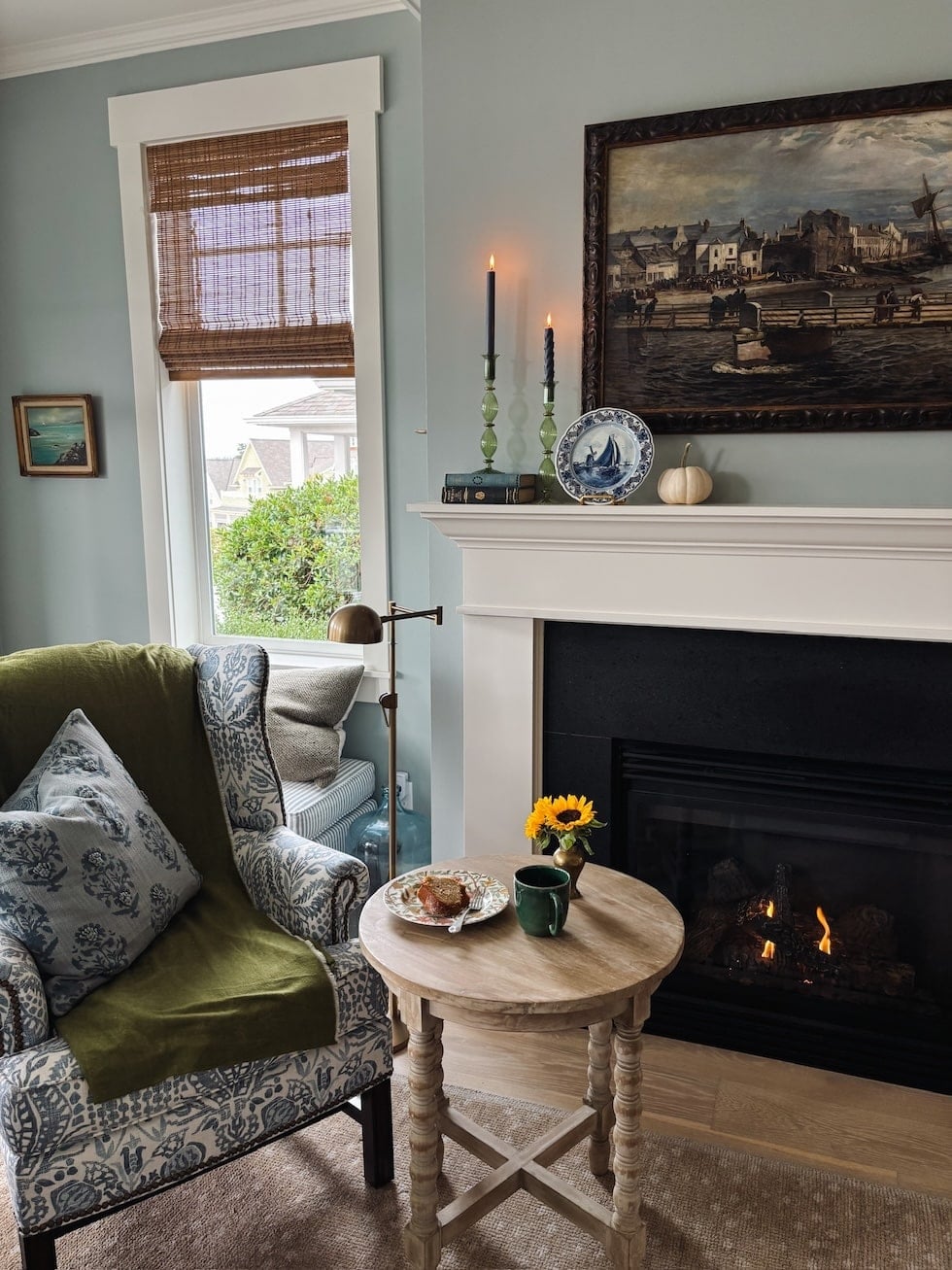 Cozy living room with green wingback chair, round wooden table, fireplace with lit logs, and framed maritime painting above white mantelpiece