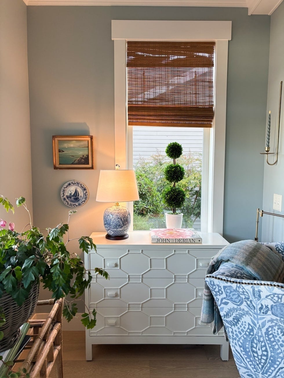 Window alcove with white wainscoting desk, blue and white ceramic plate, table lamp, potted topiary, and brown woven roman shade
